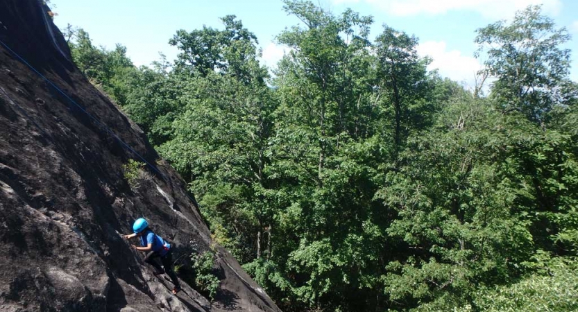 A student wearing safety gear climbs up a rock incline in front of green trees.